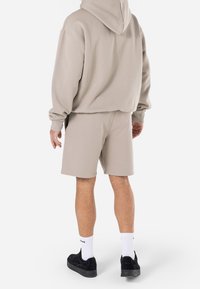 Sweat à capuche beige clair avec une poche kangourou, assorti à un short de la même couleur. Le modèle porte des baskets noires et des chaussettes blanches montantes. Tissu texturé visible.