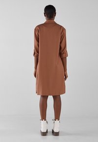 Brązowa sukienka do kolan z podwiniętymi rękawami, o prostym kroju. Noszona z białymi botkami za kostkę i gładką fakturą.