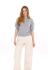 Žena s dlhými blond vlasmi, oblečená v striebornom pletenom top s krátkymi rukávmi a vysokými širokými krémovými nohavicami, stojaca na bielom pozadí.