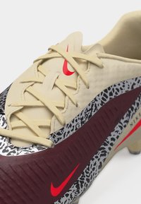 Nogometni čevelj z bež vezalkami, bordo stranskim panelom, črno-belim živalskim vzorcem in rdečim logotipom Nike Swoosh na jezičku in strani.