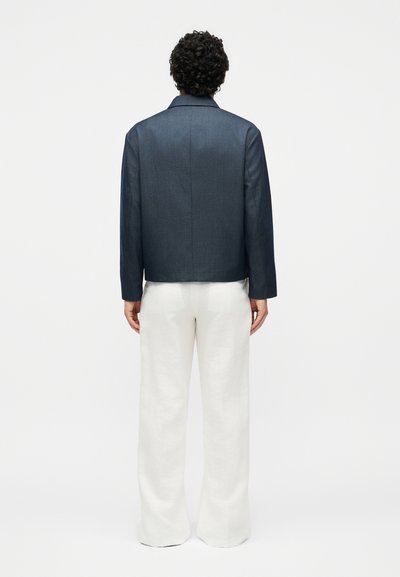 Άτομο με σγουρά σκουρόχρωμα μαλλιά που φορά μπλε σκούρο μπουφάν και φαρδιά λευκά παντελόνια, στέκεται με την πλάτη σε ένα απλό φόντο.