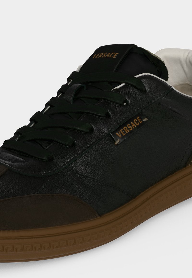 Sneaker in pelle nera con suola marrone, lacci neri e logo Versace dorato sulla linguetta e sul pannello laterale.