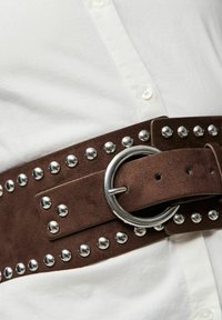 Hnědý semišový pásek se stříbrnými hroty a kruhovou kovovou sponou, v kombinaci s bílou košilí s límečkem. Texturovaný povrch s viditelným šitím.