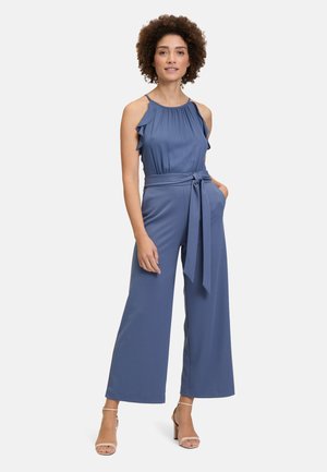 Blauwe, mouwloze jumpsuit met gerimpelde banden en een ceintuur in de taille. Vloeiende stof, ontwerp met wijd uitlopende pijpen en subtiele glans. Draagt met hakken met bandjes.