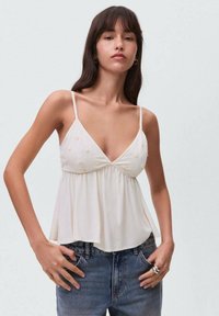 Lyse cremede cami-top med tynde stropper, der har en sammensat bærestykke og teksturerede gule blomsterapplikationer. Bæres sammen med højtoptrukne denimjeans.