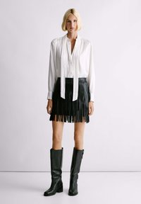 Witte blouse met lange mouwen en een strik-kraag, gecombineerd met een zwarte leren mini-jurk met franjes en zwarte leren laarzen tot de knie.