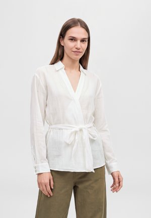 Une femme porte un chemisier blanc cache-cœur noué à la taille et un pantalon olive, sur un fond clair et uni.