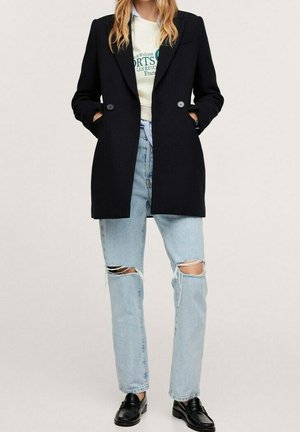 Kobieta ubrana w czarną marynarkę, białą bluzę z nadrukiem, jasnoniebieskie podarte dżinsy i czarne mokasyny, stojąca z rękami w kieszeniach.