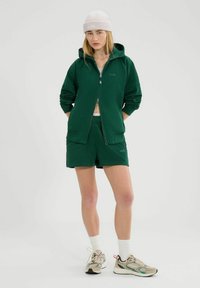 Felpa verde con cappuccio e zip frontale, abbinata a pantaloni corti coordinati. Il modello indossa calzini bianchi e sneakers beige, con un berretto chiaro.