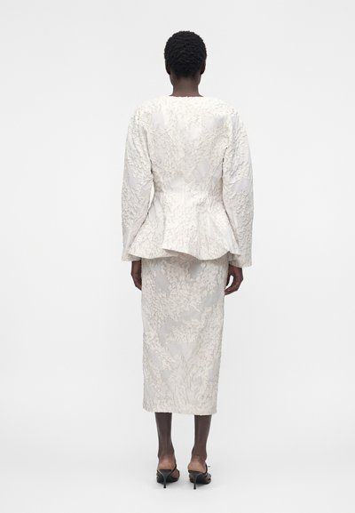 Γυναίκα φορεί ένα λευκό υφασμάτινο σακάκι με λουλουδάτο σχέδιο και ταιριαστή midi φούστα, στέκεται με την πλάτη στην κάμερα σε απλό φόντο.