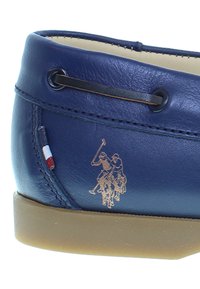 Modrá kožená bota s nízkým designem, se hnědou šňůrkou, béžovou podrážkou a zlatým emblémem zobrazujícím dva hráče póla na koni.