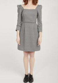 Czarna i biała sukienka w kratę z kwadratowym dekoltem, bufkami na rękawach i dopasowaną talią, sięgająca nad kolana. Noszona z czarnymi butami.