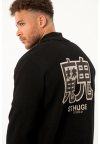 Sort uldfrakke med sjalkrave, der har en stor hvid grafik på ryggen, inklusive stiliserede tegn og teksten "STHUGE STUDIO. 21."