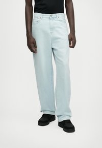 Lyseblå, vide jeans laget av denim. Har fem lommer, knappestolpe og oransje sømdetaljer. Brukes med svarte sneakers.