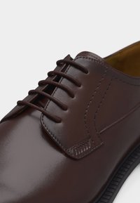 Chaussure habillée en cuir marron avec laçage fermé, finition lisse, coutures détaillées et semelle noire, vue en gros plan.
