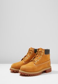 Stivali ankle in suede beige con punta rotonda, occhielli dorati, lacci marroni e un colletto nero. Suola decorata per una maggiore aderenza. Logo sul lato.