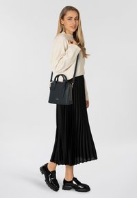 Bolso de cuero negro con asas superiores y una correa larga, acabado texturizado, herrajes dorados, combinado con un suéter de crema claro y una falda negra plisada.