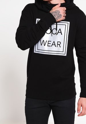 Persoon draagt een zwarte hoodie met wit vierkant logo met de tekst "sda WEAR", hand met bloemtattoo raakt nek aan, zwarte broek zichtbaar.