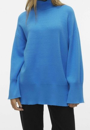 Kobieta ubrana w luźny, jasnoniebieski sweter z golfem, z długimi prążkowanymi mankietami i dołem, zestawiony z czarnymi spodniami.