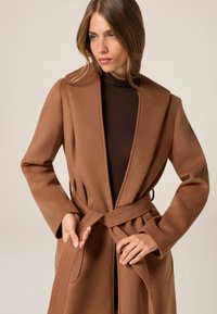 Bruine, gestructureerde wikkeljas met brede revers, een strikgordel en zijzakken, gedragen over een donkerbruine coltrui. Zachte stofafwerking.