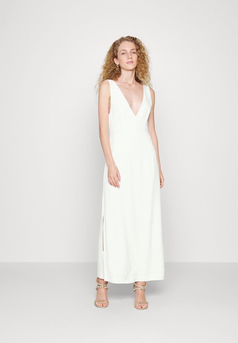 Vestido maxi branco, sem mangas, com um decote em V profundo, fendas laterais e silhueta simples. Modelo usa sandálias douradas decoradas.