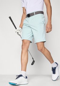 Calções de golfe azul claro com frente lisa e bolsos laterais, combinados com um cinto cinza. O conjunto inclui sapatos brancos com detalhes azuis e um taco de golfe.