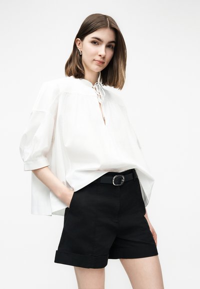 Jeune femme portant un chemisier blanc ample à manches trois-quarts et un short noir ceinturé, debout avec une main dans la poche.