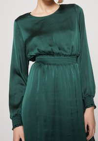 Grön satin klänning med långa ärmar, smockad midja och en mjuk, svävande design med en subtil ribbad textur. Rund halsringning.