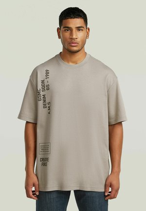 Mand stående med fronten vendt mod kamera iført en løs grå T-shirt med lodret sort tekst på venstre side og blå jeans.