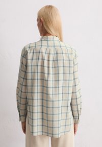 Osoba z długimi, blond włosami, ubrana w kremową i niebieską koszulę w kratę z długim rękawem oraz beżowe spodnie w prążki, stoi pod białą ścianą.