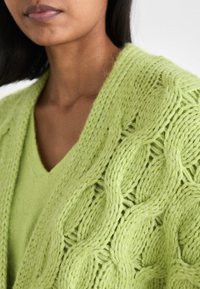Πράσινο πλεκτό ζακέτα με κυματιστή υφή και ένα βαθύ V-λαιμό από κάτω, αναδεικνύοντας ένα ζωντανό, απαλό ύφασμα.