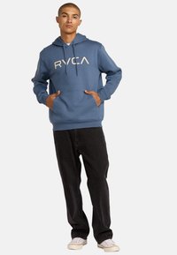 Felpa blu con grande logo beige "RYCA", tasche frontali, cappuccio con coulisse e polsini a costine, abbinata a jeans neri e sneakers bianche.