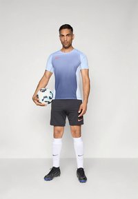 Camisola de desporto Nike com degradê azul, calções pretos, meias brancas e chuteiras de futebol pretas. Segurando uma bola de futebol com design padrão.