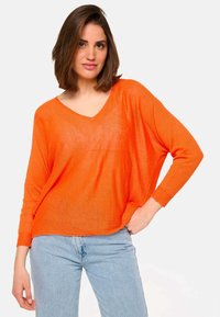 Femme avec des cheveux bruns arrivant aux épaules, portant un pull V en orange à manches longues et un jean bleu clair, se tenant avec une main dans la poche.