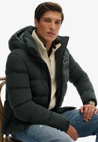 Ciemnozielona kurtka puchowa z kapturem, prążkowanym kołnierzem i zapięciem na zamek, noszona na kremowej dzianinowej bluzce. Udekorowana pikowaną podszewką i zegarkiem na nadgarstku.