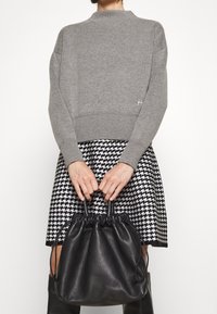 Szary sweter dzianinowy z dekoltem w serek, zestawiony z czarną i białą spódnicą w wzór houndstooth. Trzyma miękki czarny skórzany worek na sznurku.