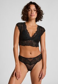 Schwarzes Spitzen-Bralette mit kurzen Ärmeln und einem tiefen V-Ausschnitt, kombiniert mit passenden durchsichtigen Spitzenunterhosen mit gepunktetem Muster und floralen Akzenten.