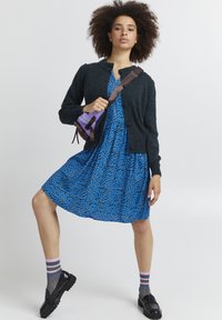 Sort cardigan, blå kjole med abstrakte mønstre, sorte sko og stribede sokker. Accessories inkluderer en lille violet taske med brune stropper.