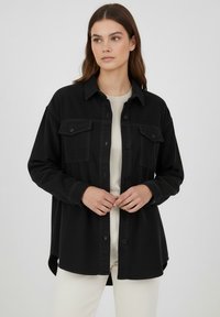 Schwarze Cord-Hemdenjacke mit Knopfleiste, zwei Brusttaschen, langen Ärmeln und abgerundetem Saum, getragen über einem hellen Oberteil.