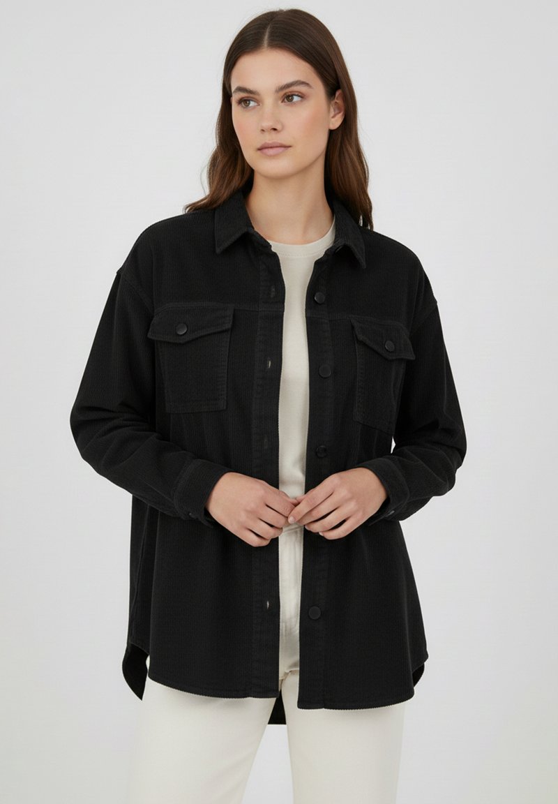 Schwarze Cord-Hemdenjacke mit Knopfleiste, zwei Brusttaschen, langen Ärmeln und abgerundetem Saum, getragen über einem hellen Oberteil.