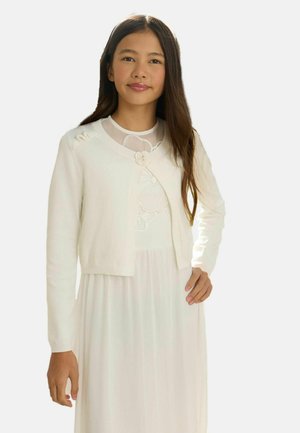 Mladé dievča s dlhými tmavými vlasmi, oblečené v bielych šatách a zodpovedajúcom bielom kardigáne s kvetinovým detailom, stojace s jednou rukou na boku.