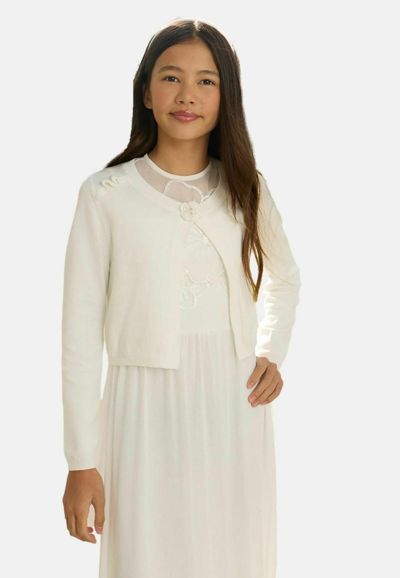 Noor tüdruk, kellel on pikk tume juuksed, valges kleidis ja selle juurde sobivas valges kardiganis lillelise detailiga, seisab ühe käe puusal.