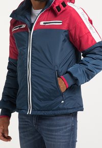 Veste rembourrée en marine et rouge, avec un col montant, une fermeture éclair à l'avant, des poches latérales et des accents blancs sur les manches. Texture douce et matelassée.