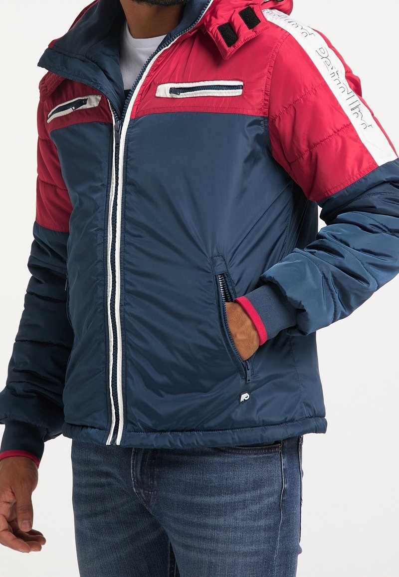 Veste rembourrée en marine et rouge, avec un col montant, une fermeture éclair à l'avant, des poches latérales et des accents blancs sur les manches. Texture douce et matelassée.