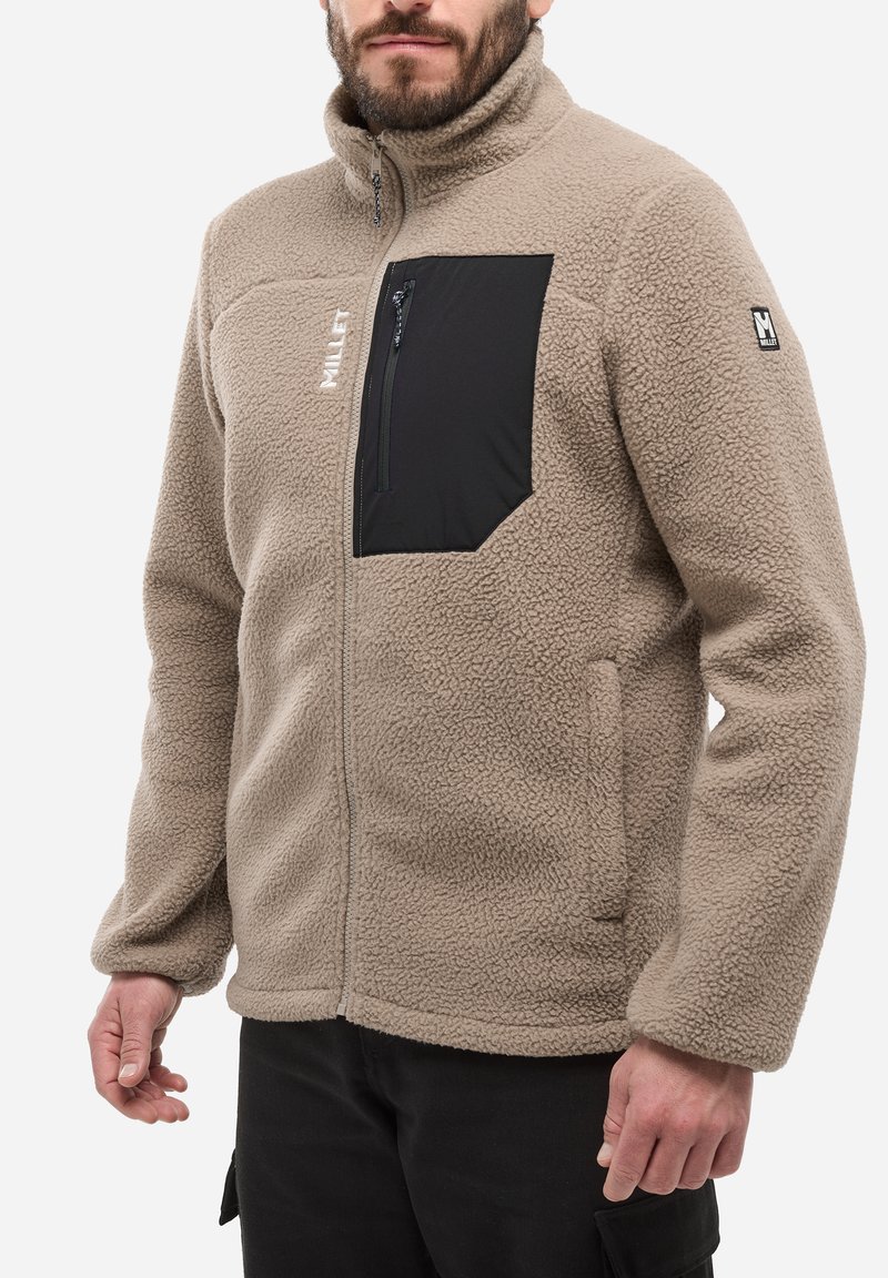 Homme portant une veste en polaire beige avec une poche poitrine zippée noire et un logo "Millet" blanc, assortie à un pantalon noir.