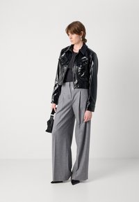 Fekete lakk, rövid fazonú dzseki gombos és cipzáros záródással, szürke, bő szárú nadrágok, hegyes orrú fekete magassarkú cipők és kis kézitáska. Egyszerű háttér.