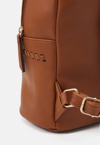 Sac à dos en cuir marron avec une texture lisse, matériel doré et détails de couture décoratifs sur le côté. Sangle ajustable incluse.