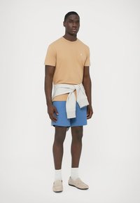 Bež majica kratkih rukava, plave kratke hlače, sivi džemper vezan oko struka, bež mokasinke, bijele čarape; jednostavan dizajn bez uzoraka.