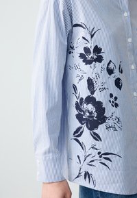 Camicia a righe in blu chiaro e bianco, con stampa floreale scura su un lato. Maniche lunghe con polsini abbottonati. Tessuto in cotone.