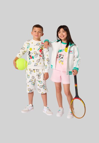 Ragazzo e ragazza vestiti con abiti a tema tennis di Topolino, che tengono una pallina da tennis e una racchetta, sorridenti davanti a uno sfondo grigio.
